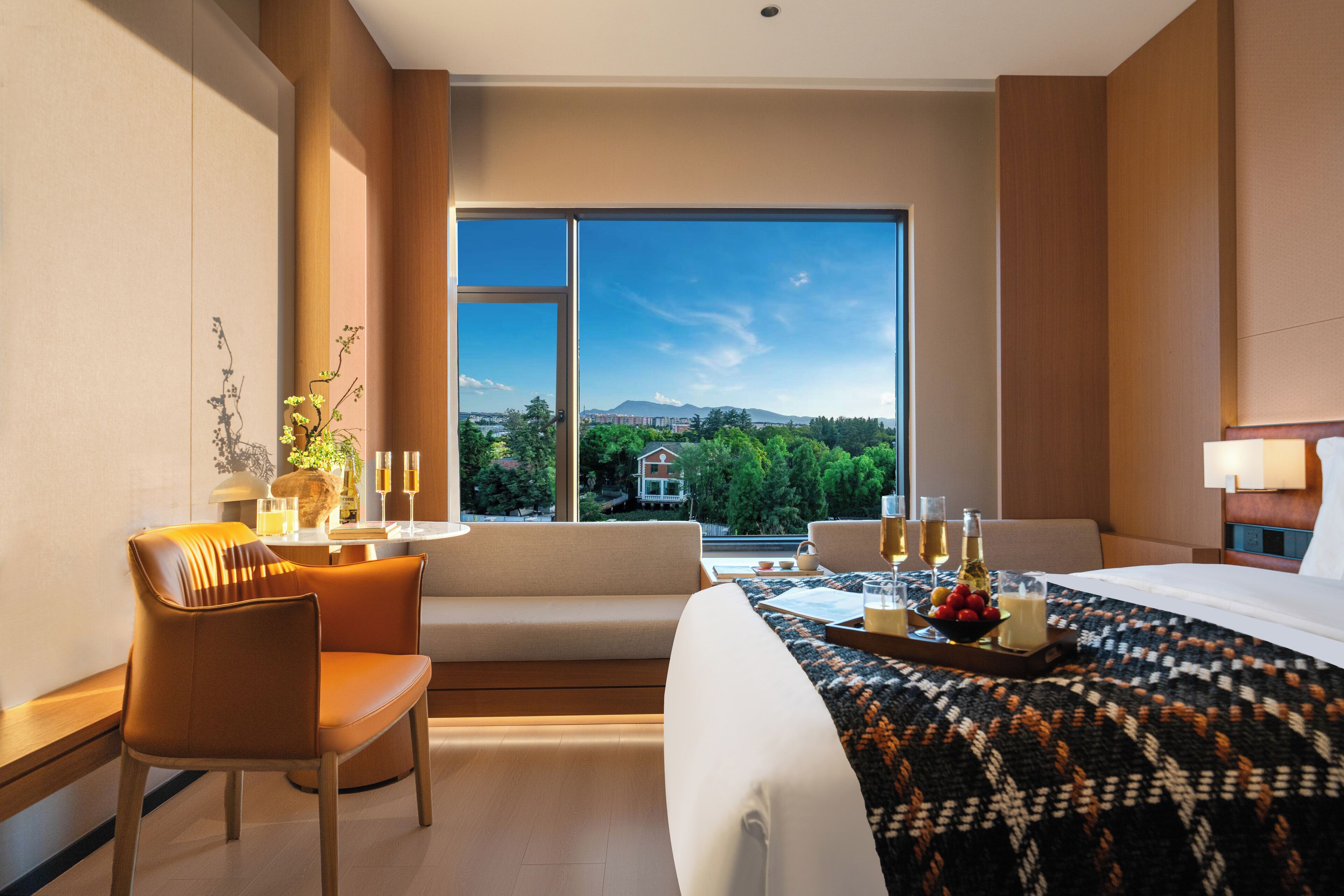 A hotel room with a large window, a sofa, a bed, and a dining area with a tray of fruit and drinks.
