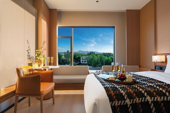 Habitación de hotel con un ventanal grande, sofá, cama y zona de comedor con una bandeja de frutas y bebidas.