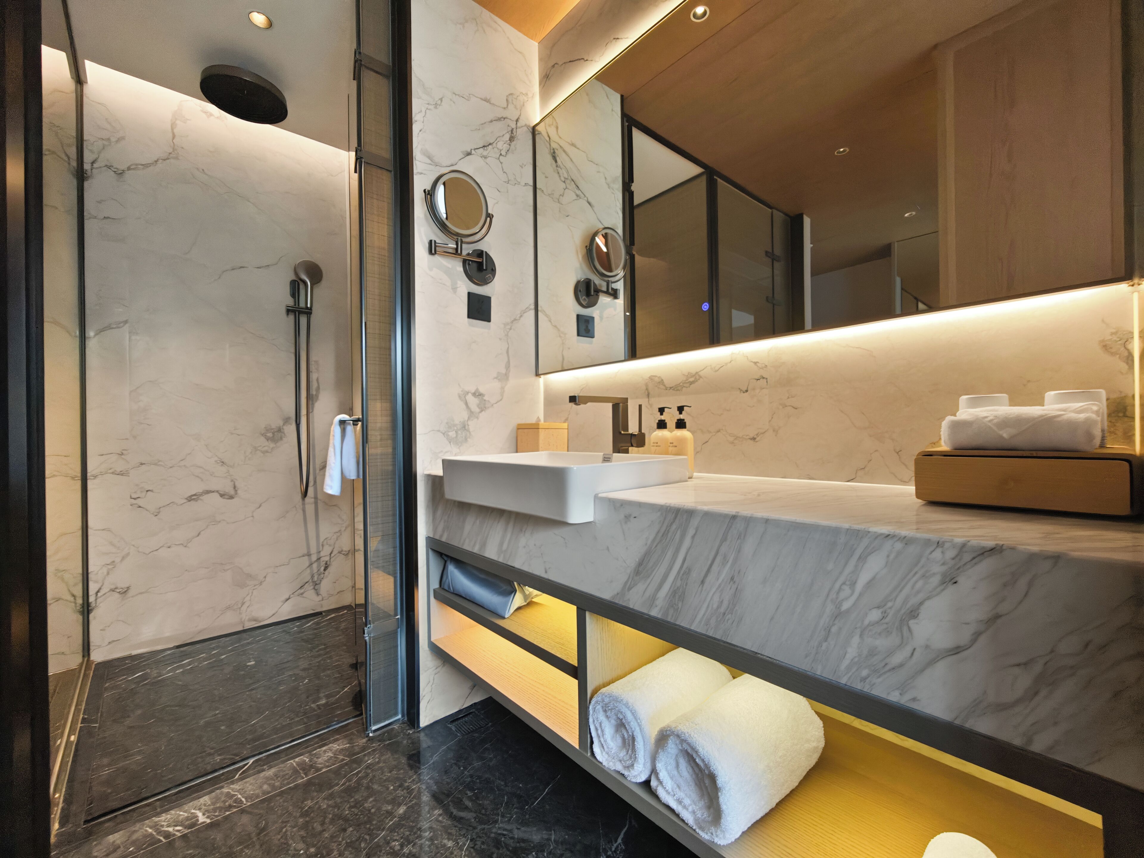 A modern bathroom with a glass shower enclosure, marble walls, a large mirror, and a wooden vanity with a towel rack.