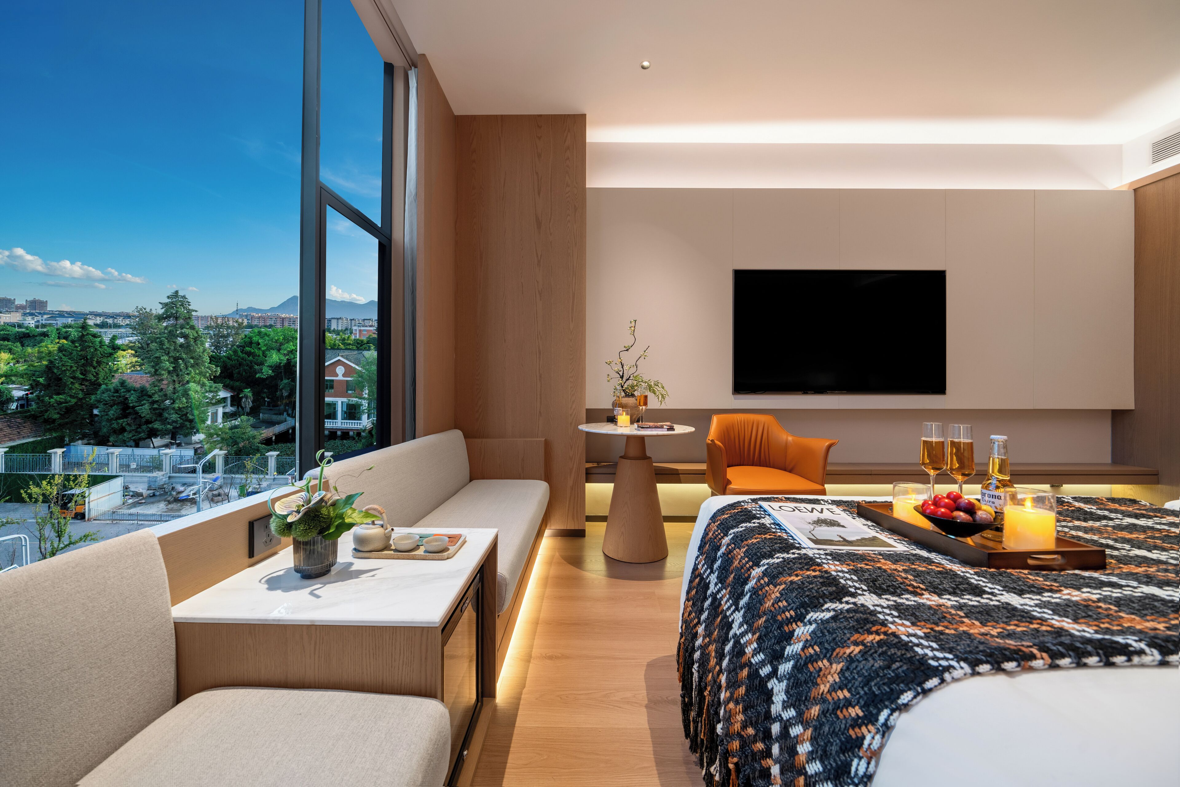 Une chambre d’hôtel moderne dotée d’une grande fenêtre, d’un canapé, d’un lit, d’une petite table avec une plante et d’un plateau avec des boissons et des fruits.