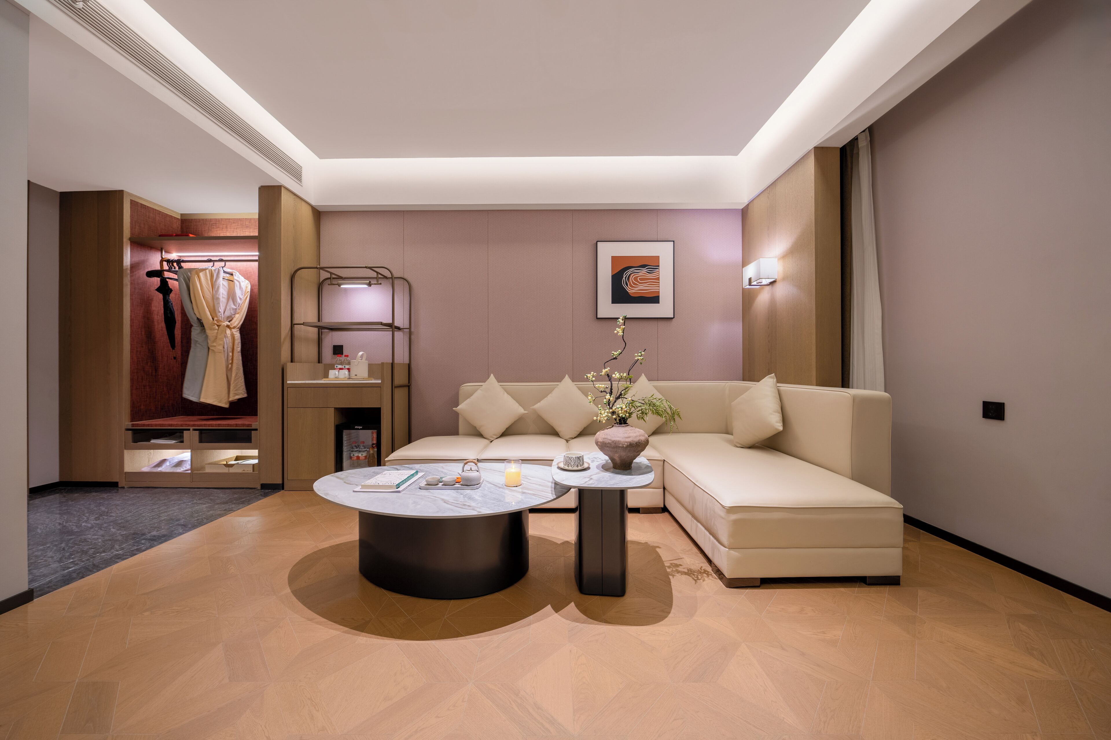 A modern living room with a sofa, a coffee table, and a small side table. There is a coat rack with clothes and a shelf with decorative items.