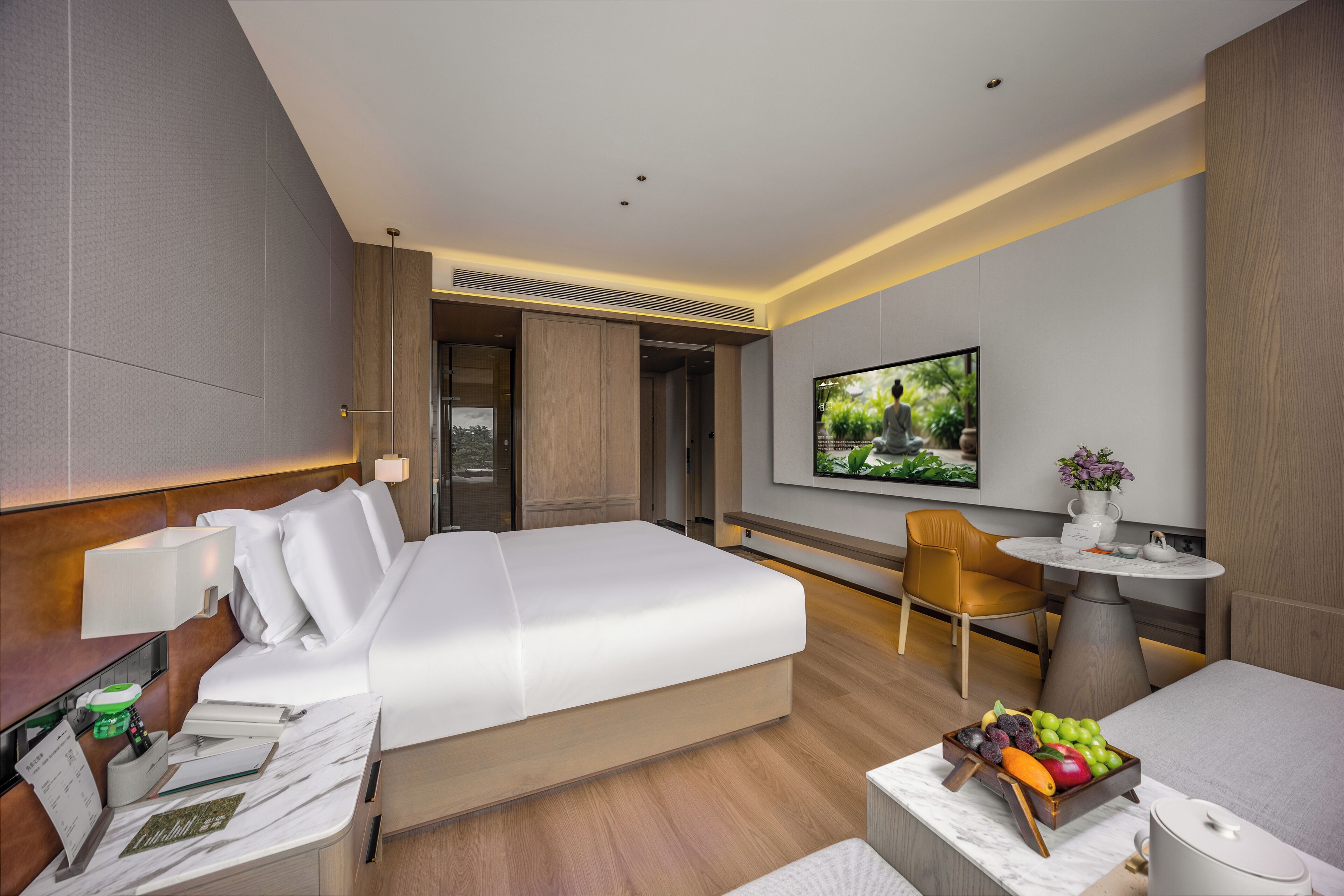 A modern hotel room with a large bed, a desk with a chair, a small table with a vase, and a fruit bowl.