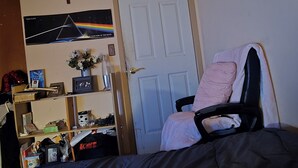A bedroom with a bed, a chair, a shelf with various items, a door, and a wall with a cross and a picture. - no longer here (Suffern)