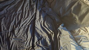 A bed with a grey blanket and pillows, a plain wall, and a partial view of a window with curtains. - no longer here (Suffern)