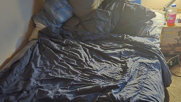A bed with a dark blue comforter and pillows, a lamp on a bedside table, and a box with a floral design. - no longer here (Suffern)