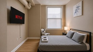 A bedroom with a bed, a TV mounted on the wall, a chair, a window with blinds, and a painting on the wall. - Playhouse SQ Downtown, R&R Hall, Clinic, CSU (Cleveland)