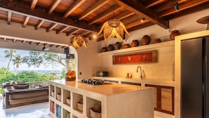 A modern kitchen with a central island, wooden ceiling beams, and a view of palm trees. - Infinity Pool | Five Bedrooms | Beachfront Stay (Canggu)