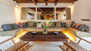A modern living room with a wooden ceiling, a large sofa with multiple cushions, a coffee table with a vase of flowers, and two side tables. - Infinity Pool | Five Bedrooms | Beachfront Stay (Canggu)