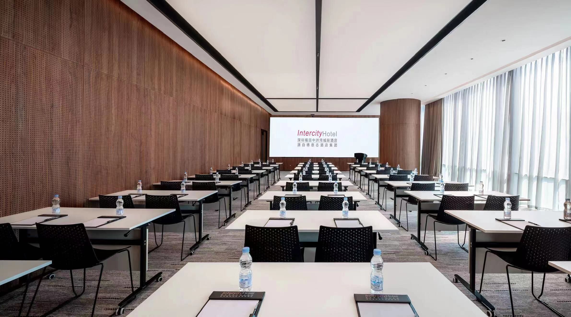 A conference room with rows of tables and chairs, a large screen at the front, and a wooden wall.