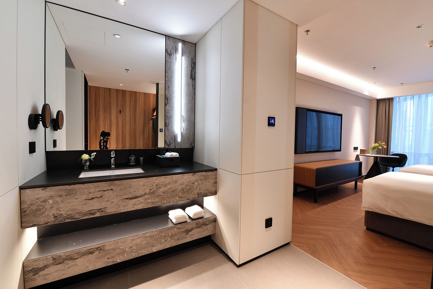 Una habitación de hotel moderna con un espejo grande, un tocador de madera, un televisor de pantalla plana, una cama y una zona de estar.