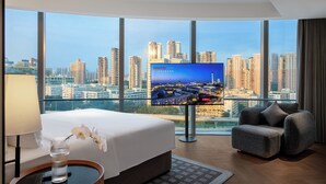 Habitación de hotel con una televisión grande, una cama, un sofá, una mesita de vidrio y vistas a la ciudad.