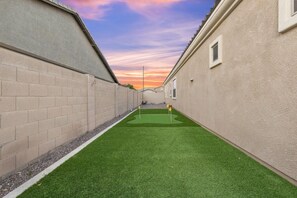 A narrow alley with artificial green turf, beige walls, and a sunset sky. - WoodHaven, Amazing Interior, Stunning Backyard, Combined Pool & Spa 11 x 20 Feet (Goodyear)