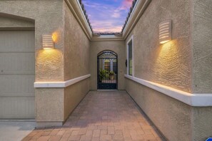 Une allée étroite mène à une entrée fermée par un portail, surmontée d’une arche décorative, et bordée de murs à motifs et de lampes fixées aux murs.