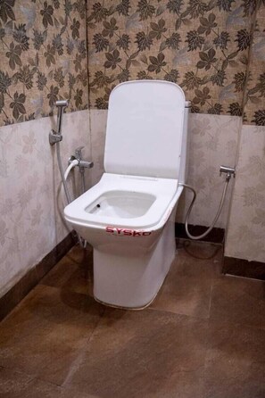 A white bidet with a raised seat and a hose attachment, set against a wall with a floral pattern. - Hotel Bombay Central Agra (Agra)