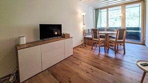 Una habitación acogedora con televisión en un estante de madera, una mesa de comedor con sillas y ventanas grandes con cortinas.
