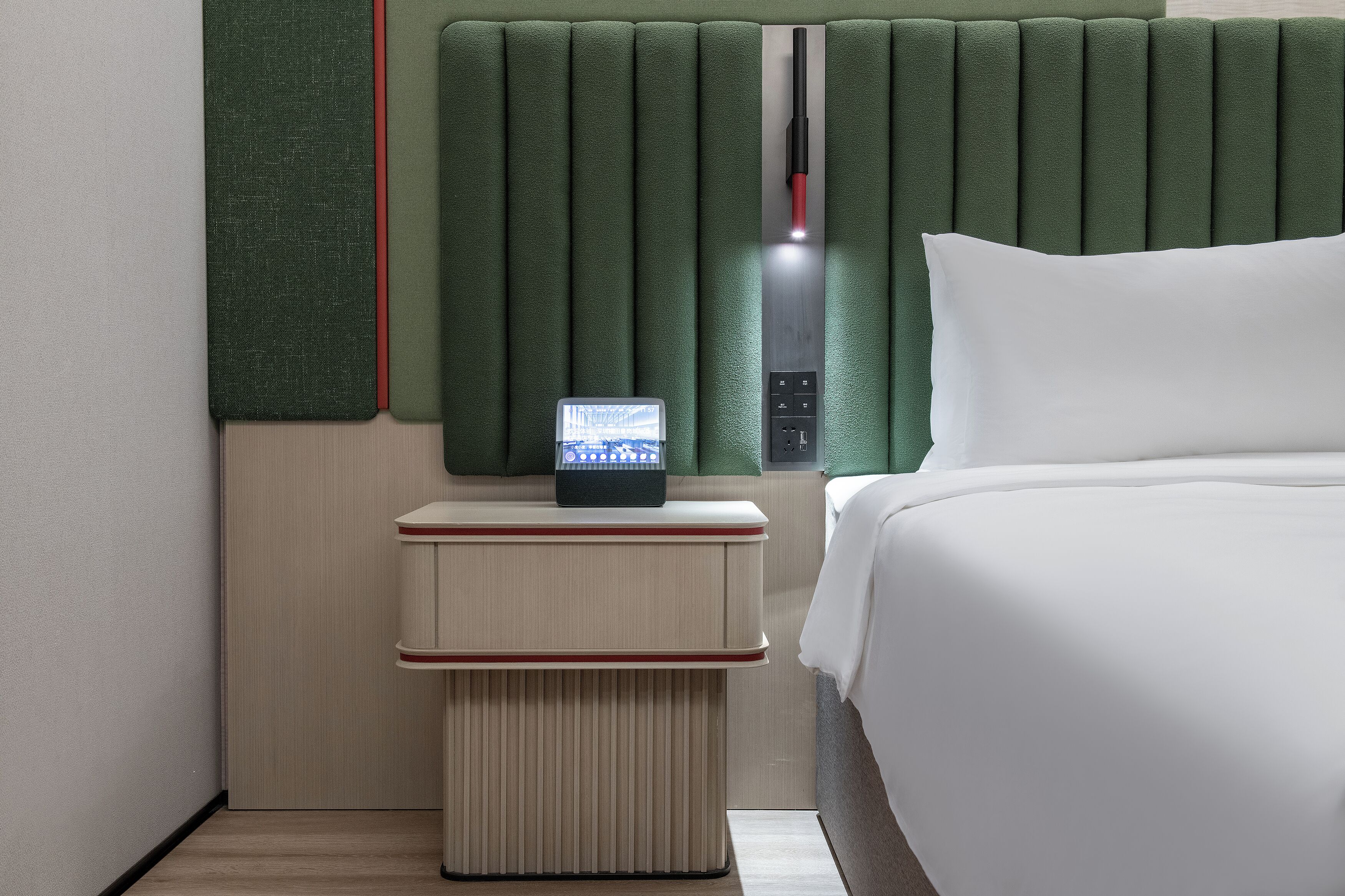 A modern hotel room with a bed, a nightstand with a digital clock, and a green headboard.