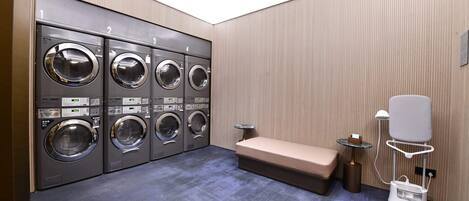 A laundry room with four washing machines, a bench, a small table with a lamp, and a hairdryer.