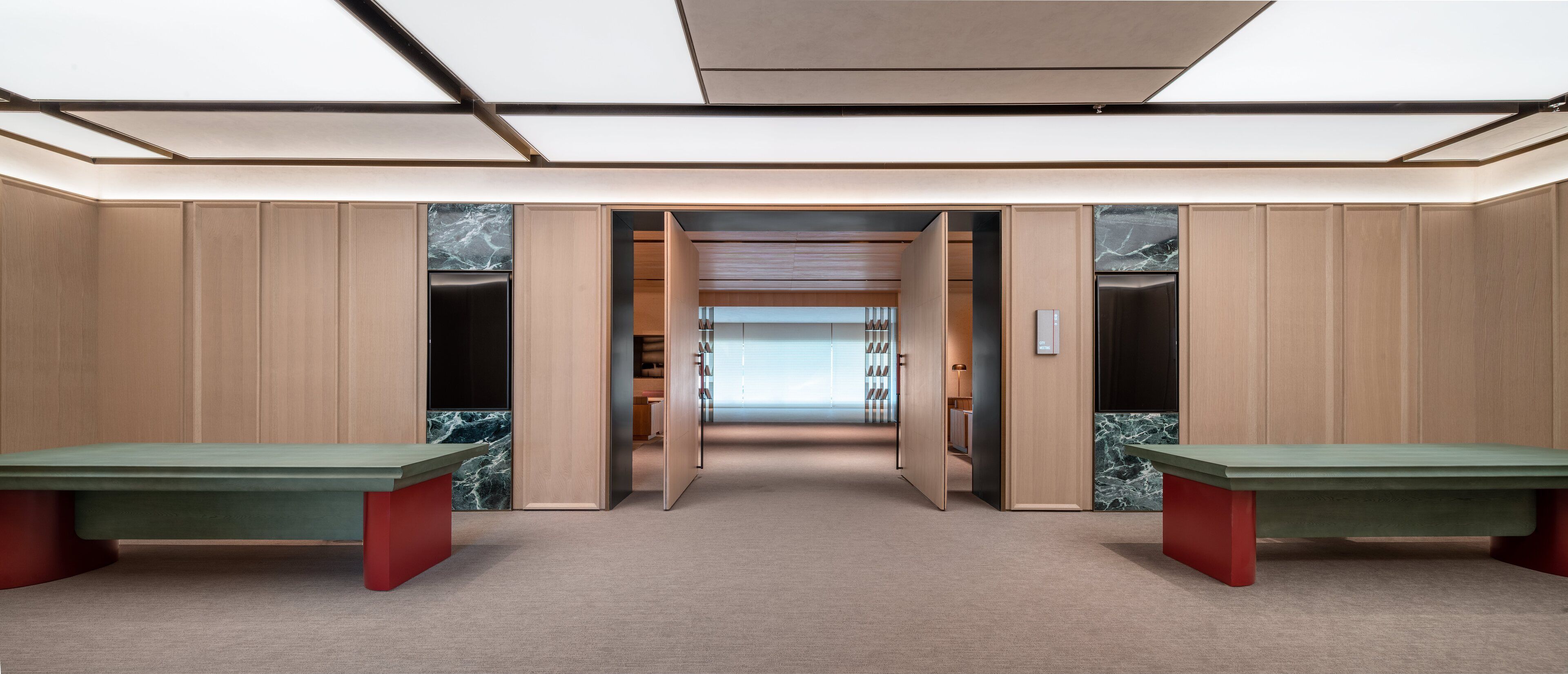 A modern, minimalist room with two green benches, wooden paneling, and a view into a library.