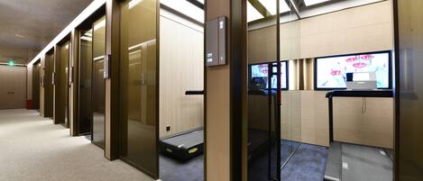 Un área de ascensores moderna con puertas de vidrio, una cinta caminadora y una pantalla digital.