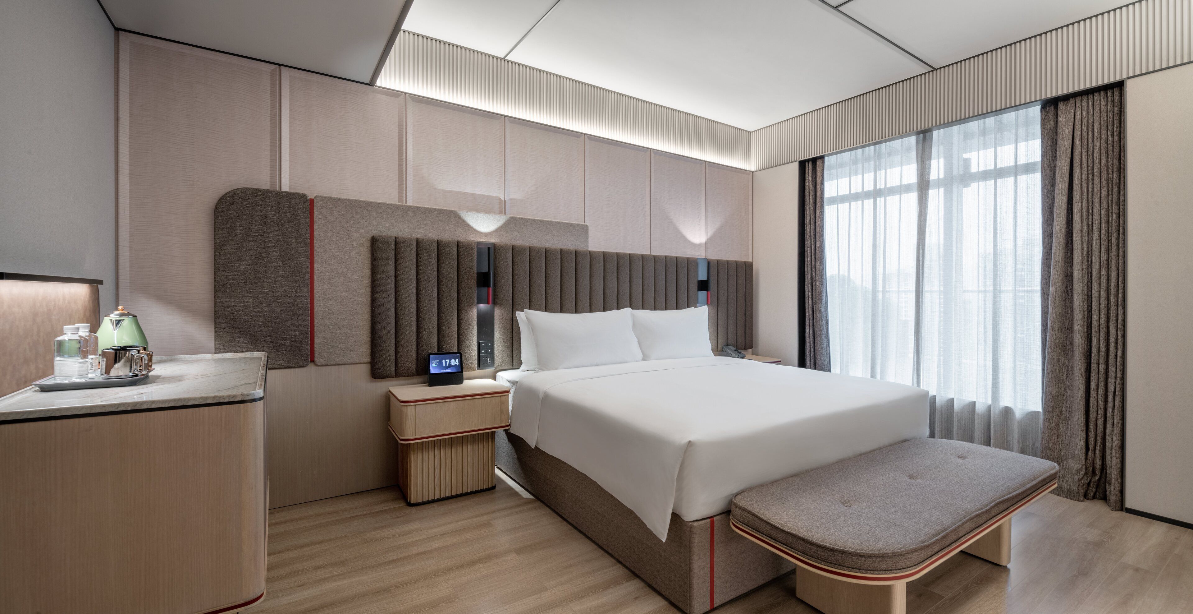 A modern hotel room with a large bed, bedside tables, a small desk with a kettle, and a window with curtains.
