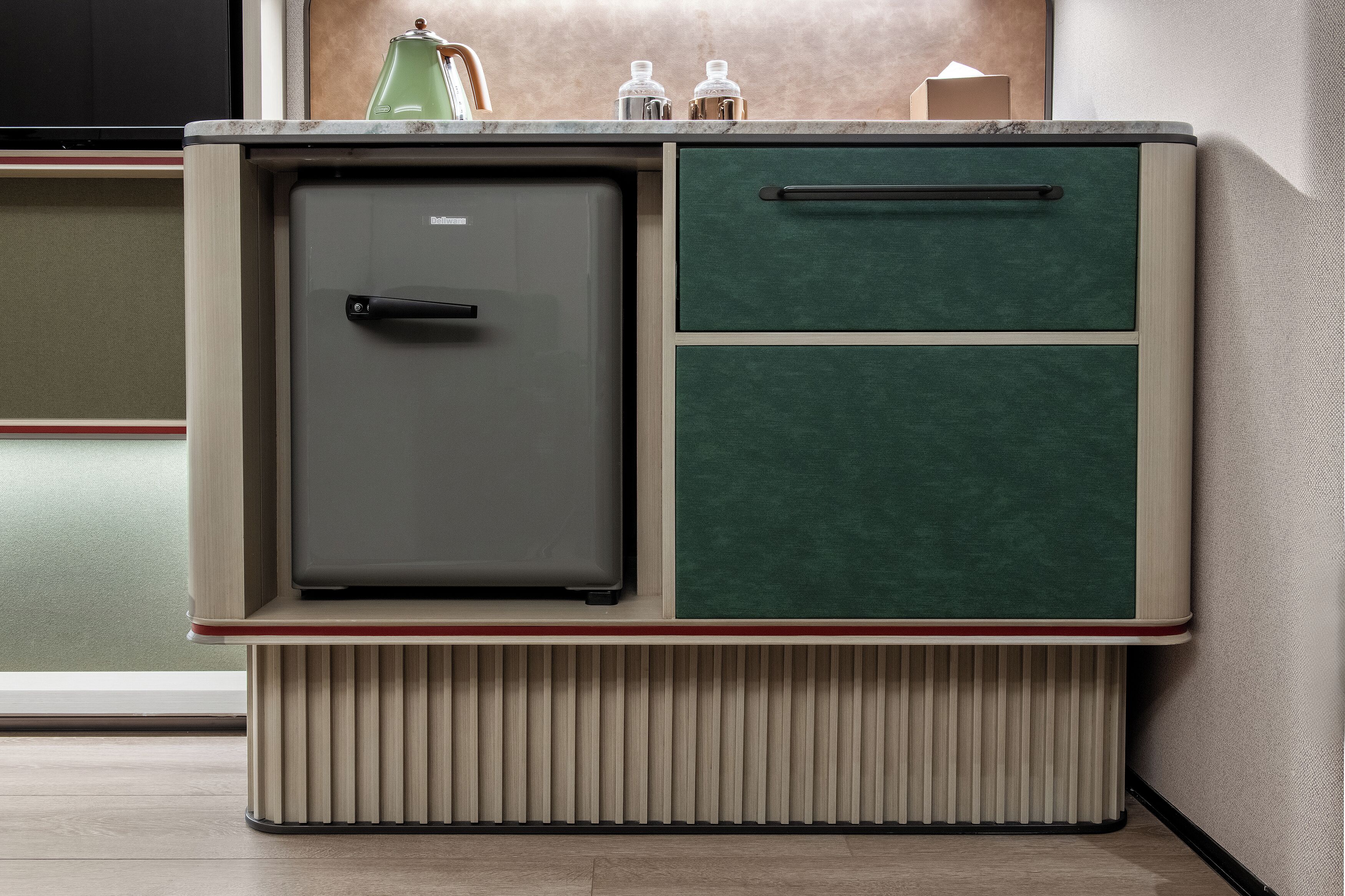 A compact kitchenette with a built-in refrigerator, a single drawer, and a countertop with a kettle and containers.