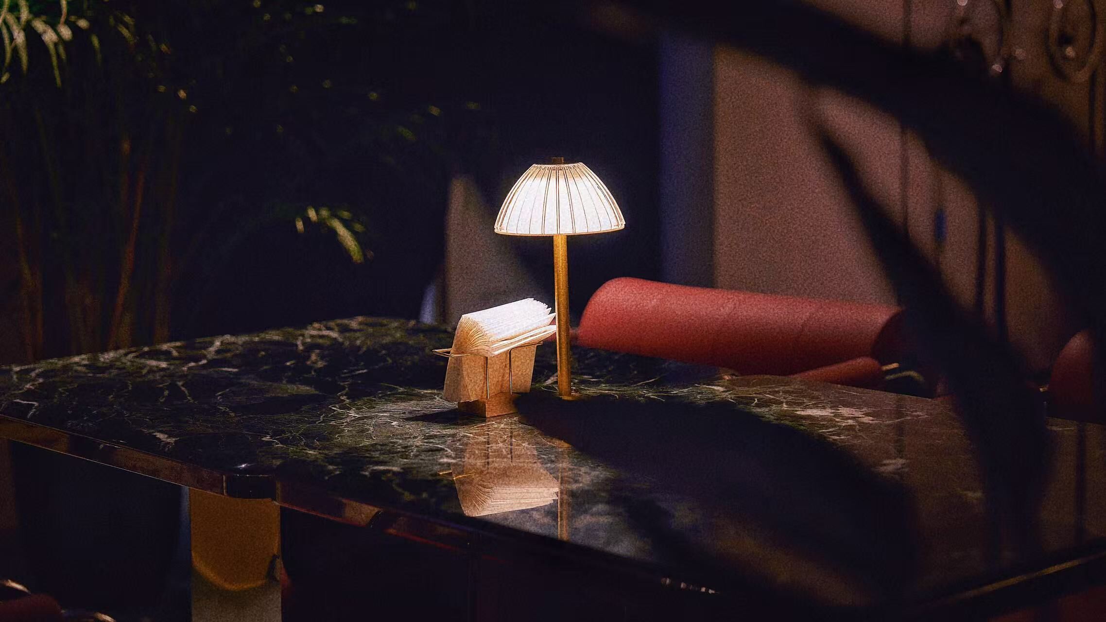 Una mesa con una lámpara, un libro y una silla roja en una habitación con poca luz.