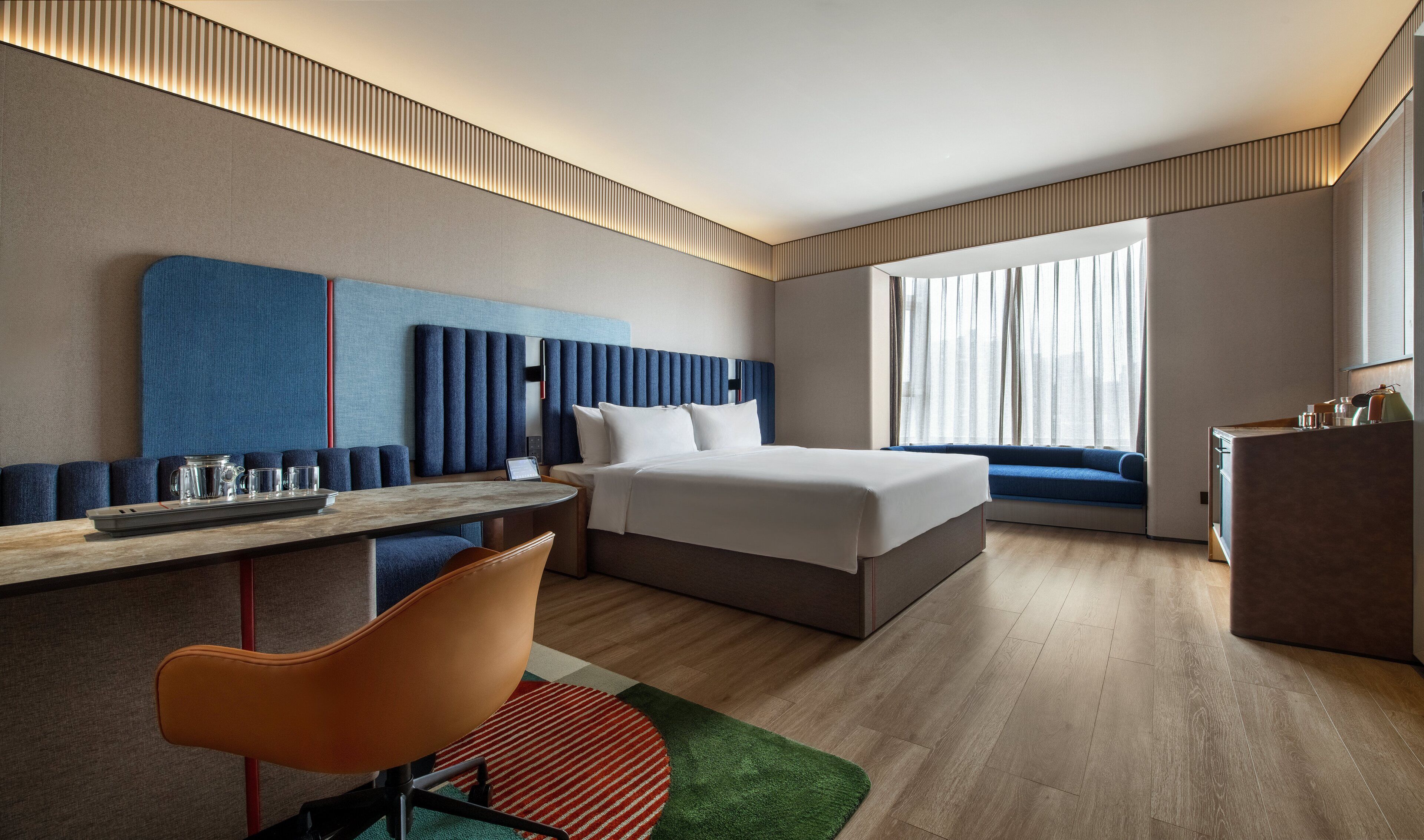 Una habitación de hotel moderna con cama, zona de estar, escritorio y vistas de la ciudad a través de amplias ventanas.