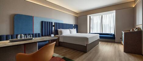 Una habitación de hotel moderna con cama, zona de estar, escritorio y vistas de la ciudad a través de amplias ventanas.