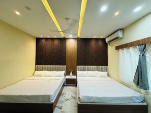 Habitación de hotel con dos camas, ventilador de techo, iluminación empotrada, pared de paneles de madera, unidad de aire acondicionado y ventana con cortinas.