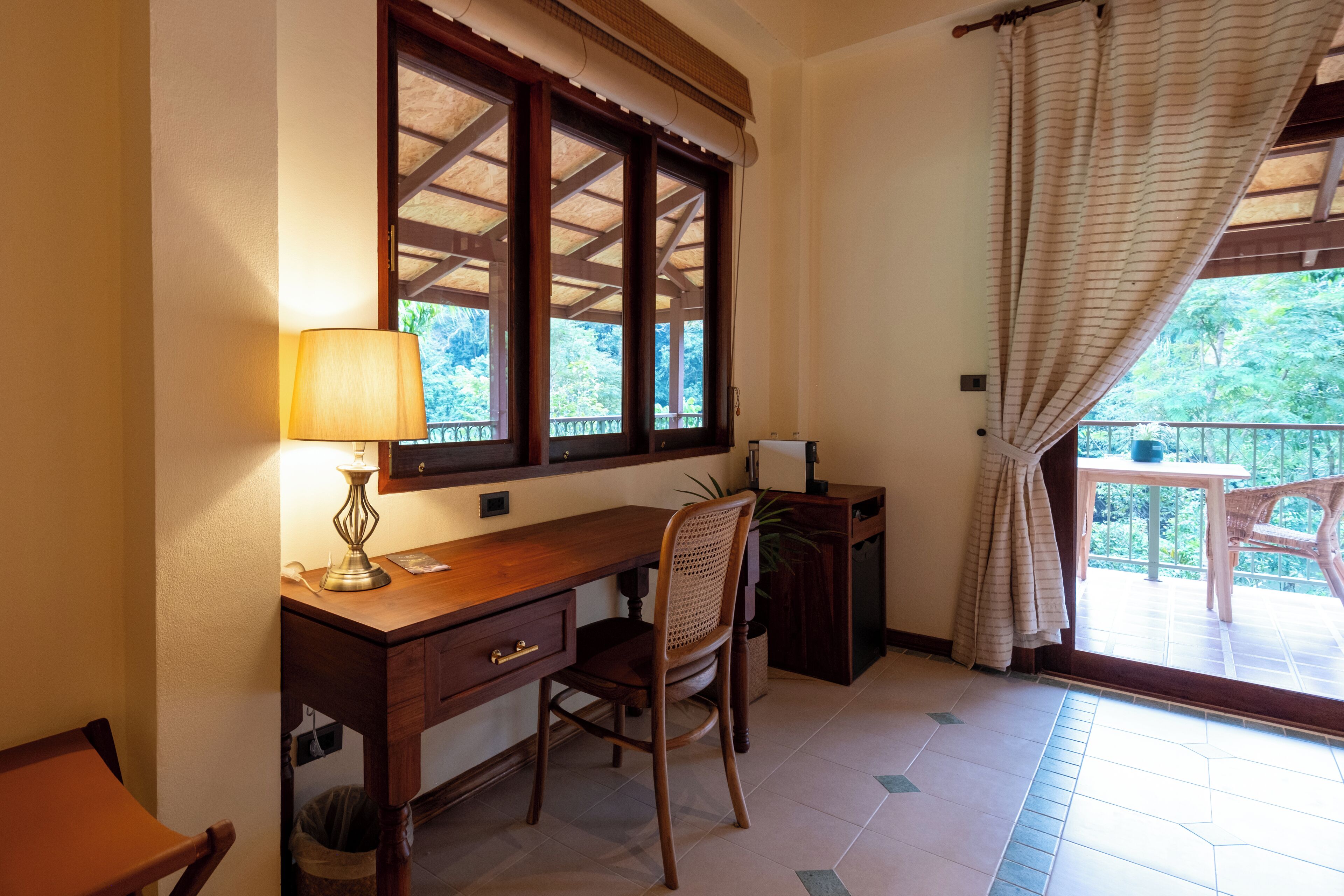 Una habitación con un escritorio de madera, una silla, una lámpara, un ventanal con cortinas y vistas al exterior.