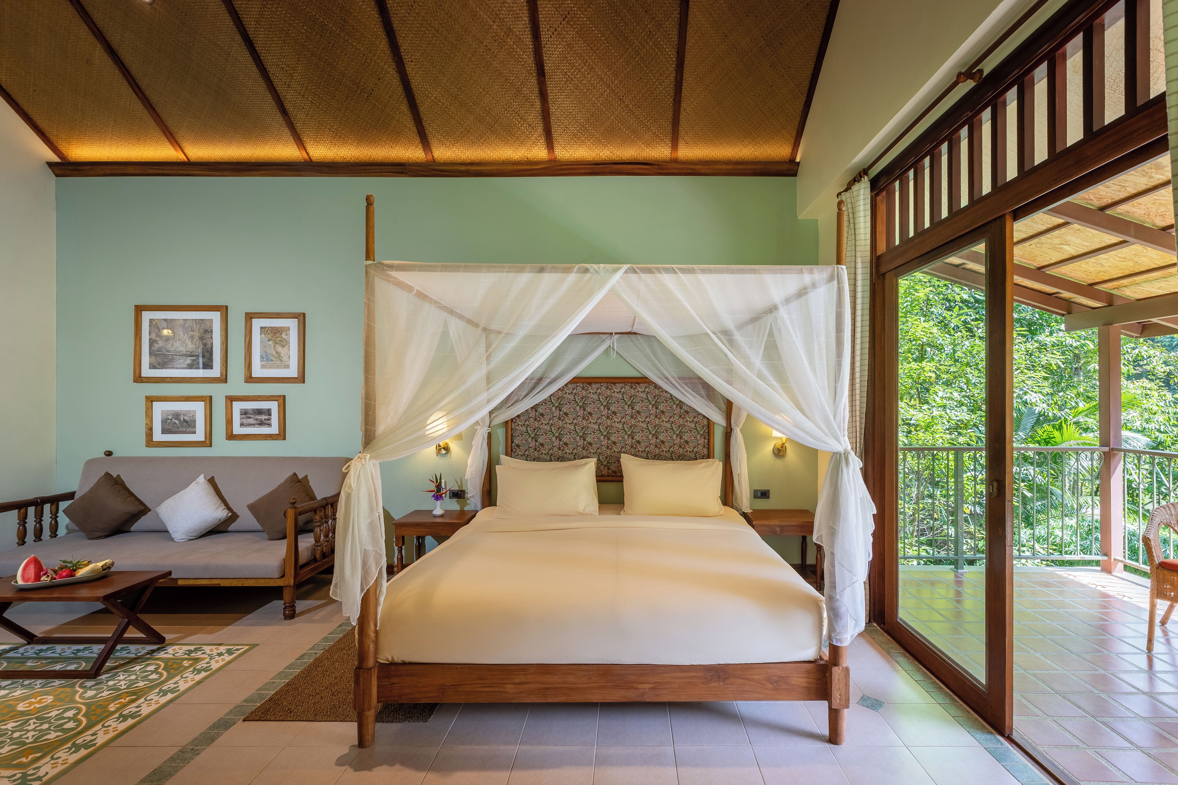 A bedroom with a four-poster bed, a sofa, a nightstand, and a large window with a view of greenery.
