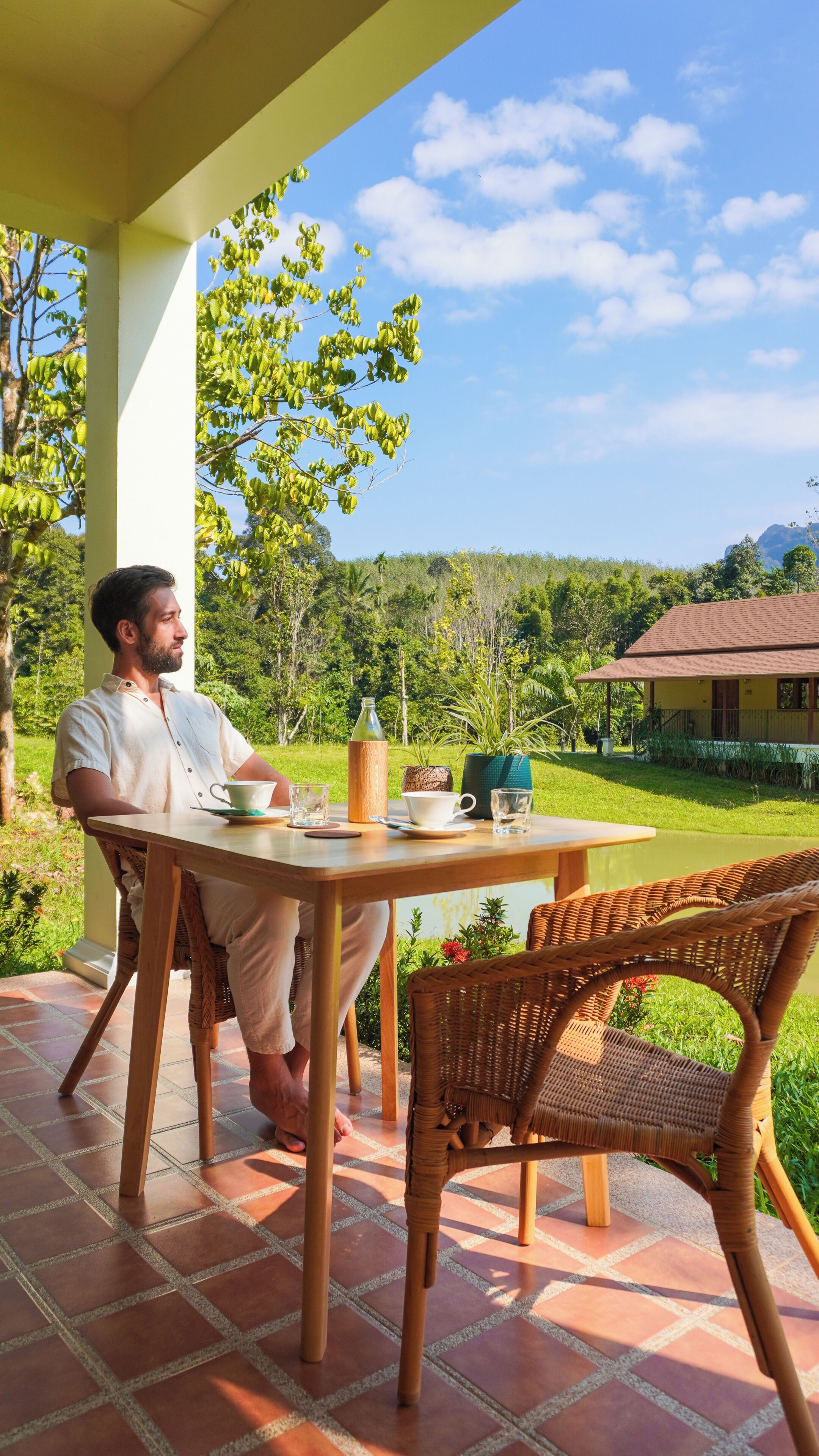 A man sitting at a table with a cup and saucer, a bottle, and a glass on a patio with a view of a house and trees.