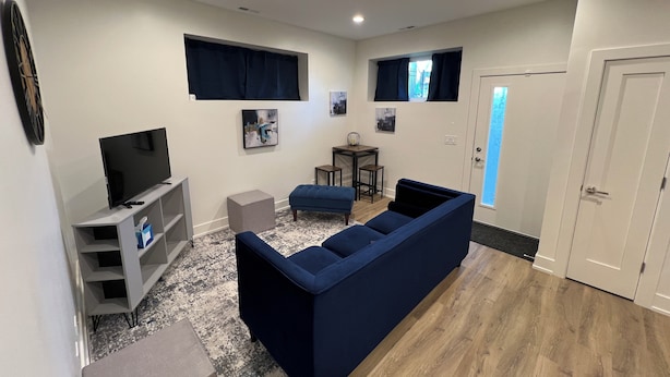 A modern living room with a sectional sofa, a TV on a stand, a small dining area, and a door leading to another room.