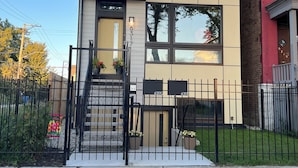 A modern two-story house with a beige and white facade, a black metal fence, and a small front yard with grass and a potted plant.
