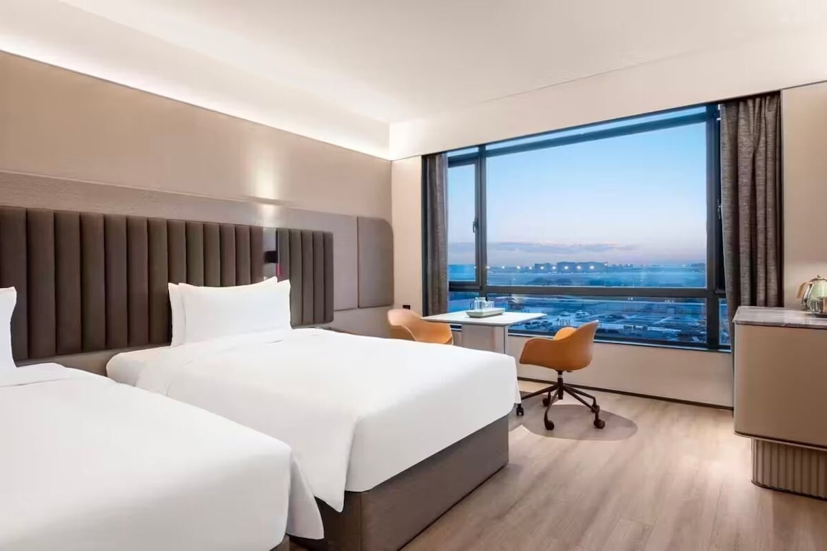 A hotel room with two beds, a desk, a chair, and a large window with a city view.