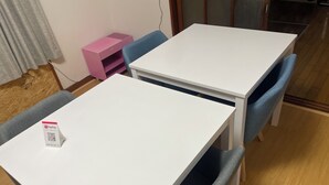 Um quarto pequeno e bem organizado, com duas mesas brancas e cadeiras azuis. Uma mesinha lateral rosa é visível no canto.