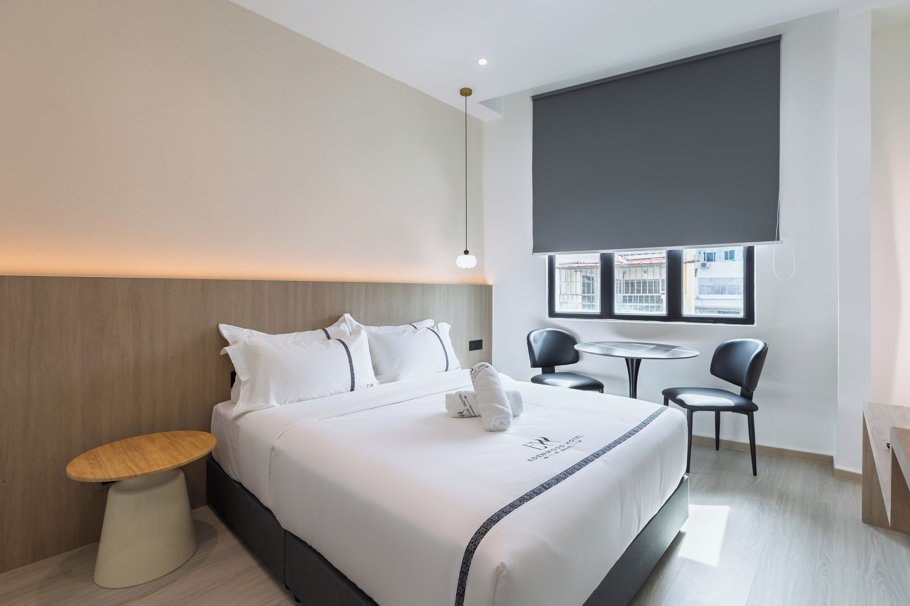 A modern hotel room with a neatly made bed, a small round table, two chairs, and a large window with a view of buildings.