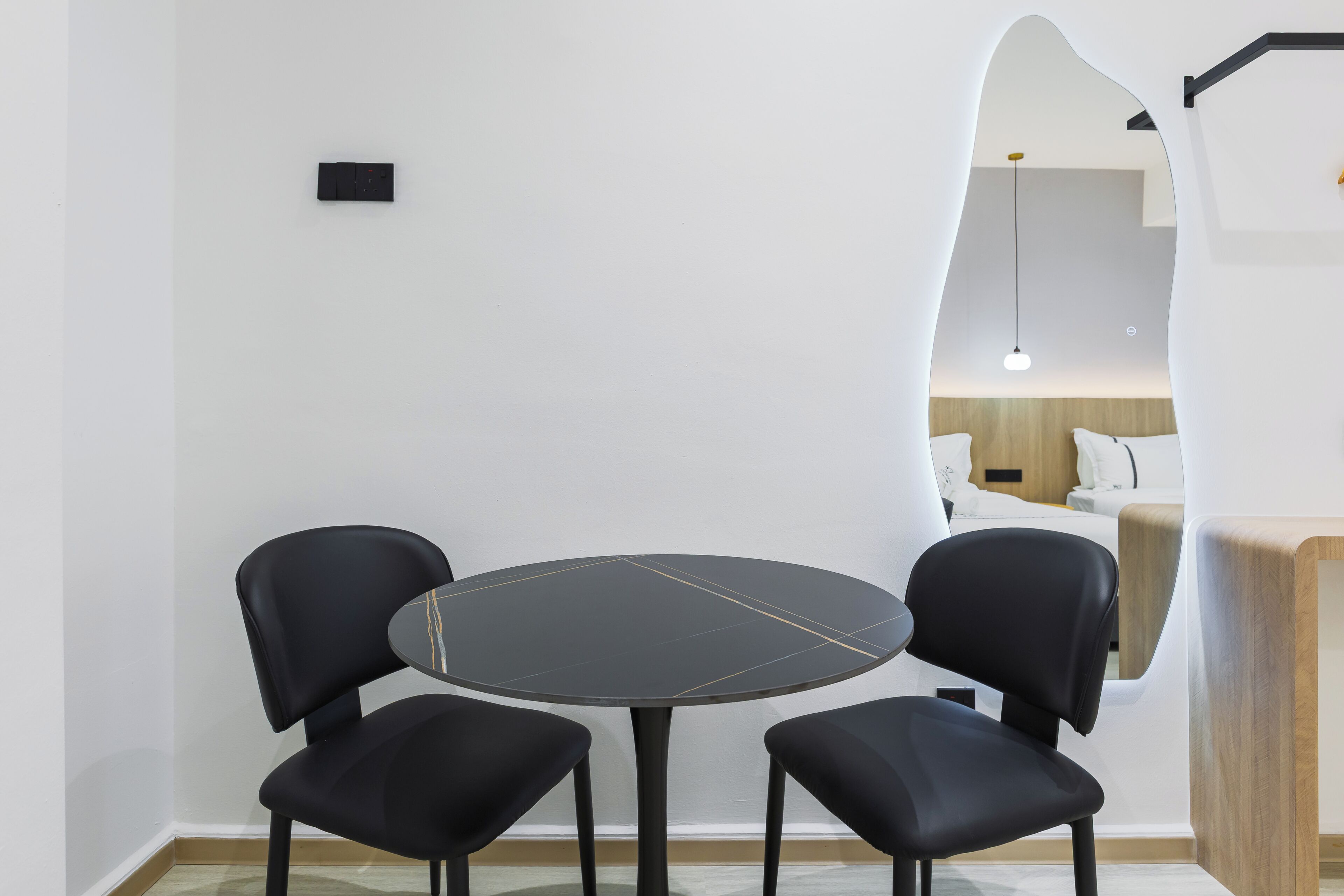 Ein modernes Zimmer mit einem runden Tisch, zwei schwarzen Stühlen, einer Holzkommode und einem Spiegel, der ein Bett und eine Pendelleuchte reflektiert.