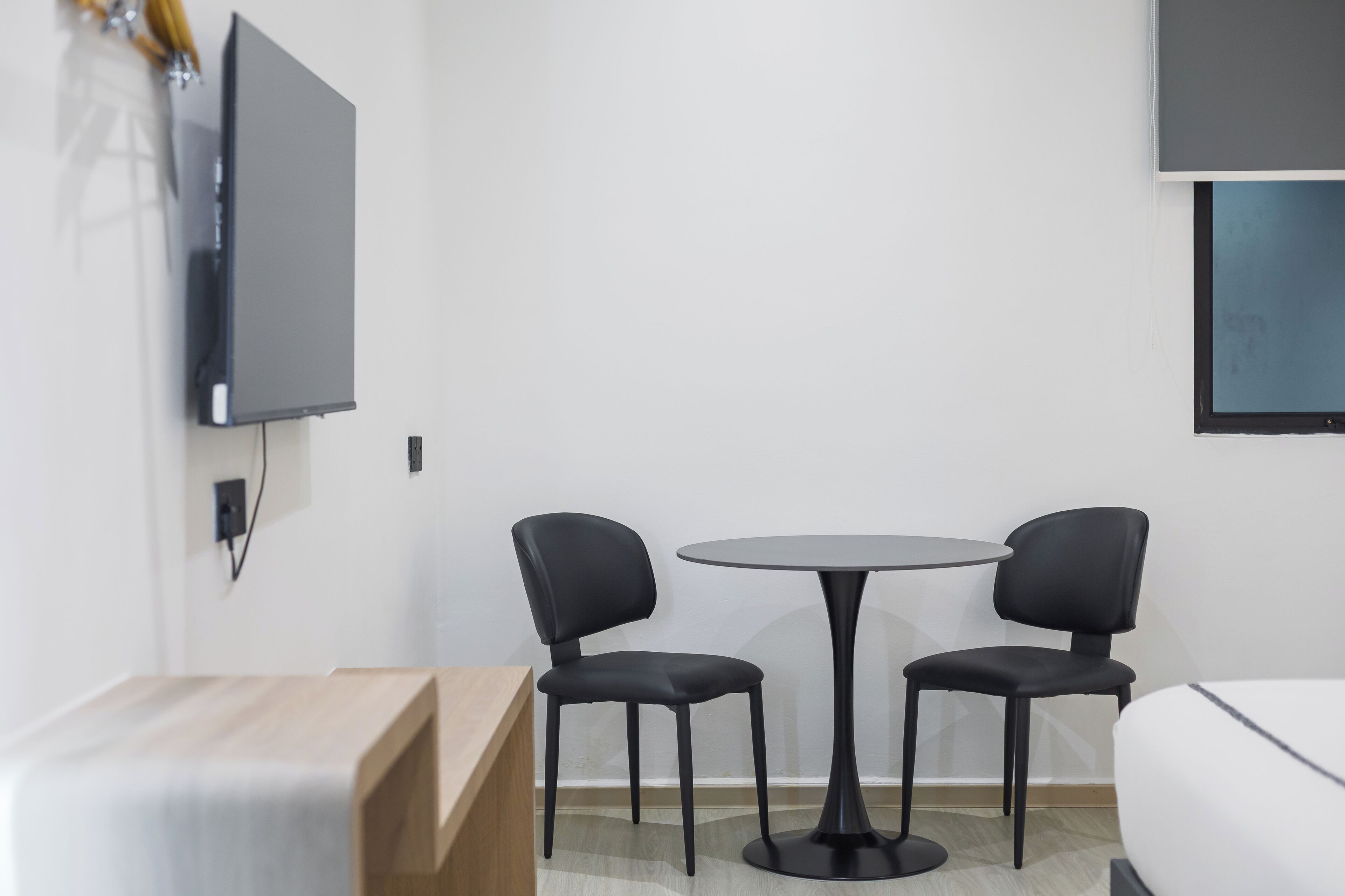 A modern hotel room with a wooden desk, two black chairs, a round table, a flat-screen TV mounted on the wall, and a bed.