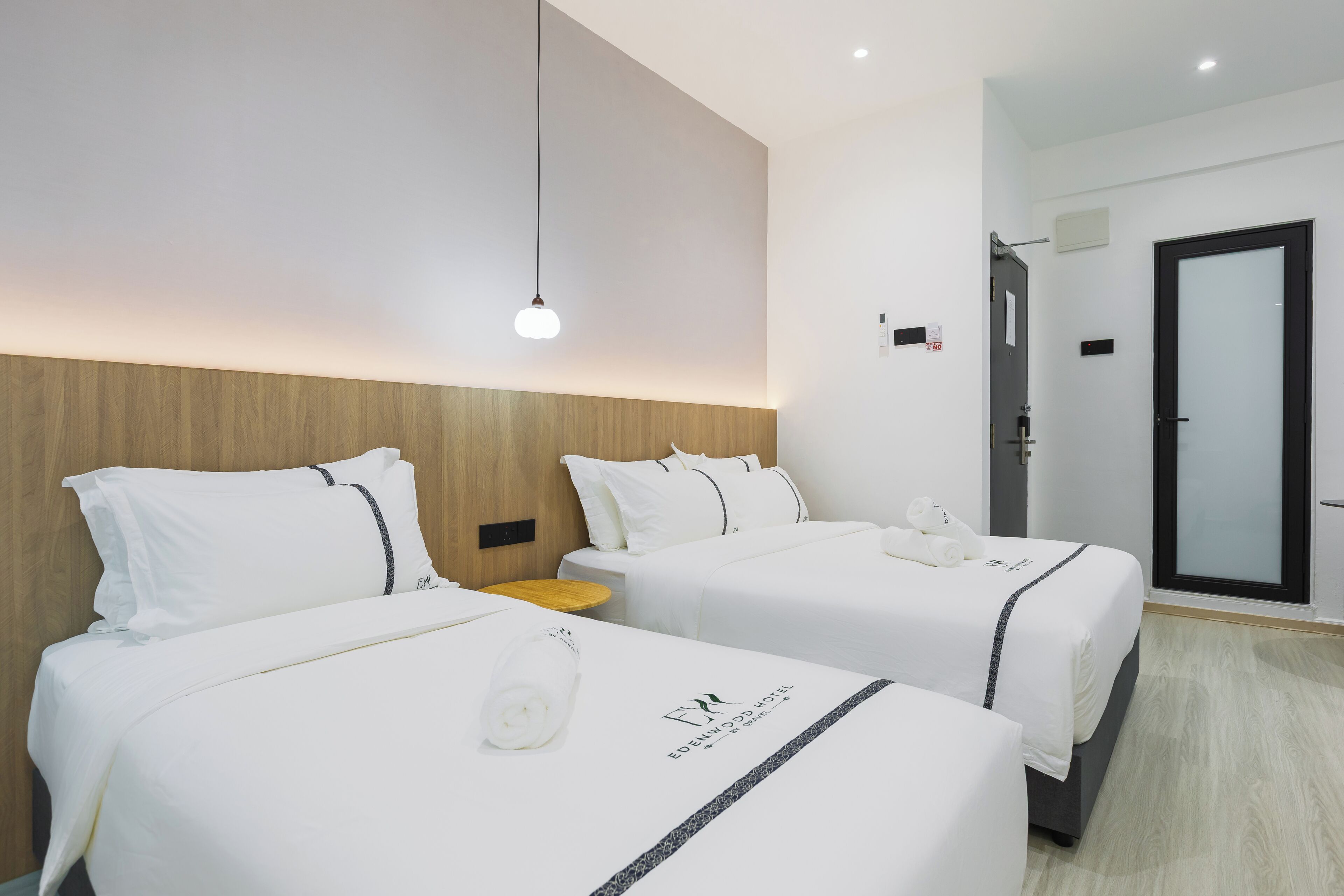 A hotel room with two beds, a wooden headboard, a small wooden side table, and a glass door leading to a bathroom.