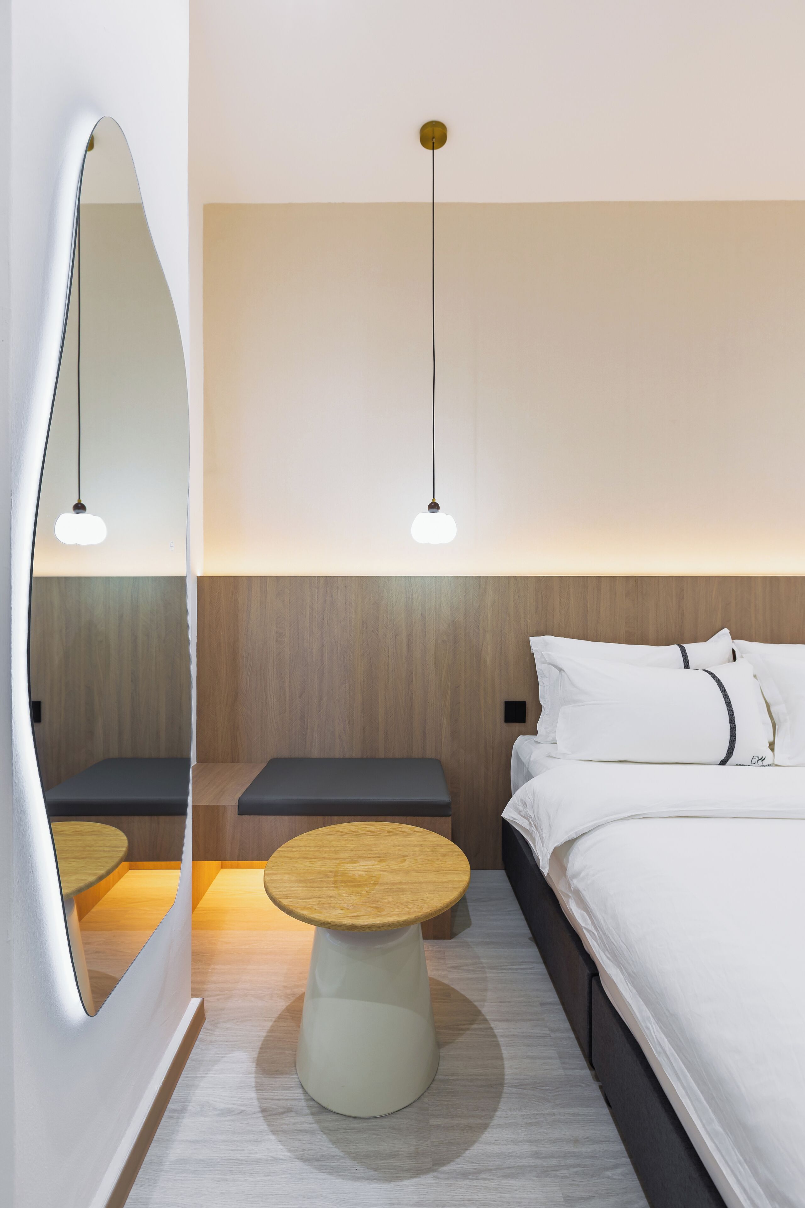 A modern hotel room with a large bed, a bedside table, a small stool, and a mirror.