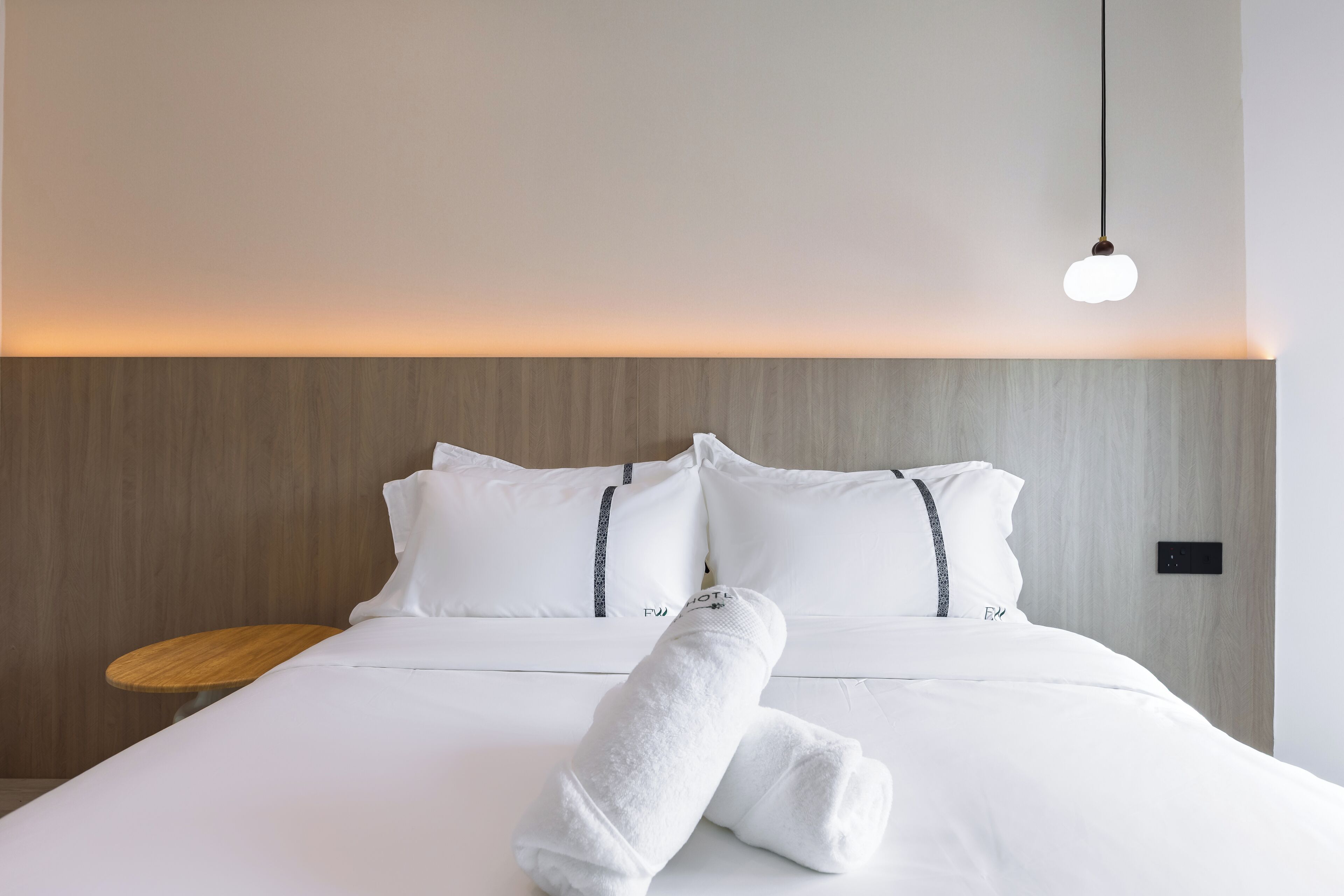 Un lit bien fait, avec du linge de lit blanc et une serviette pliée, une table de chevet avec un plateau en bois et un luminaire suspendu.
