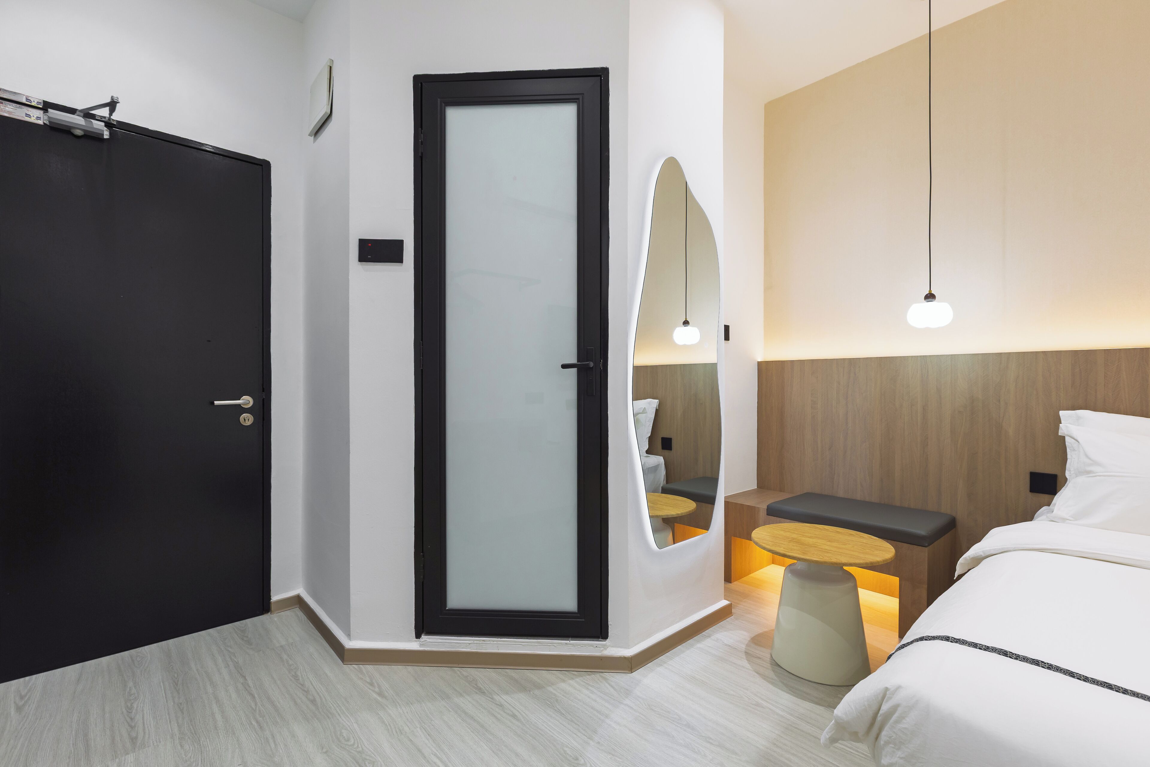 A modern hotel room with a black door, a glass partition, a bed with white bedding, a small round table with a bench, and a wall-mounted mirror.