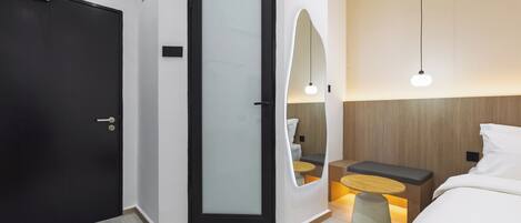 A modern hotel room with a black door, a glass partition, a bed with white bedding, a small round table with a bench, and a wall-mounted mirror.