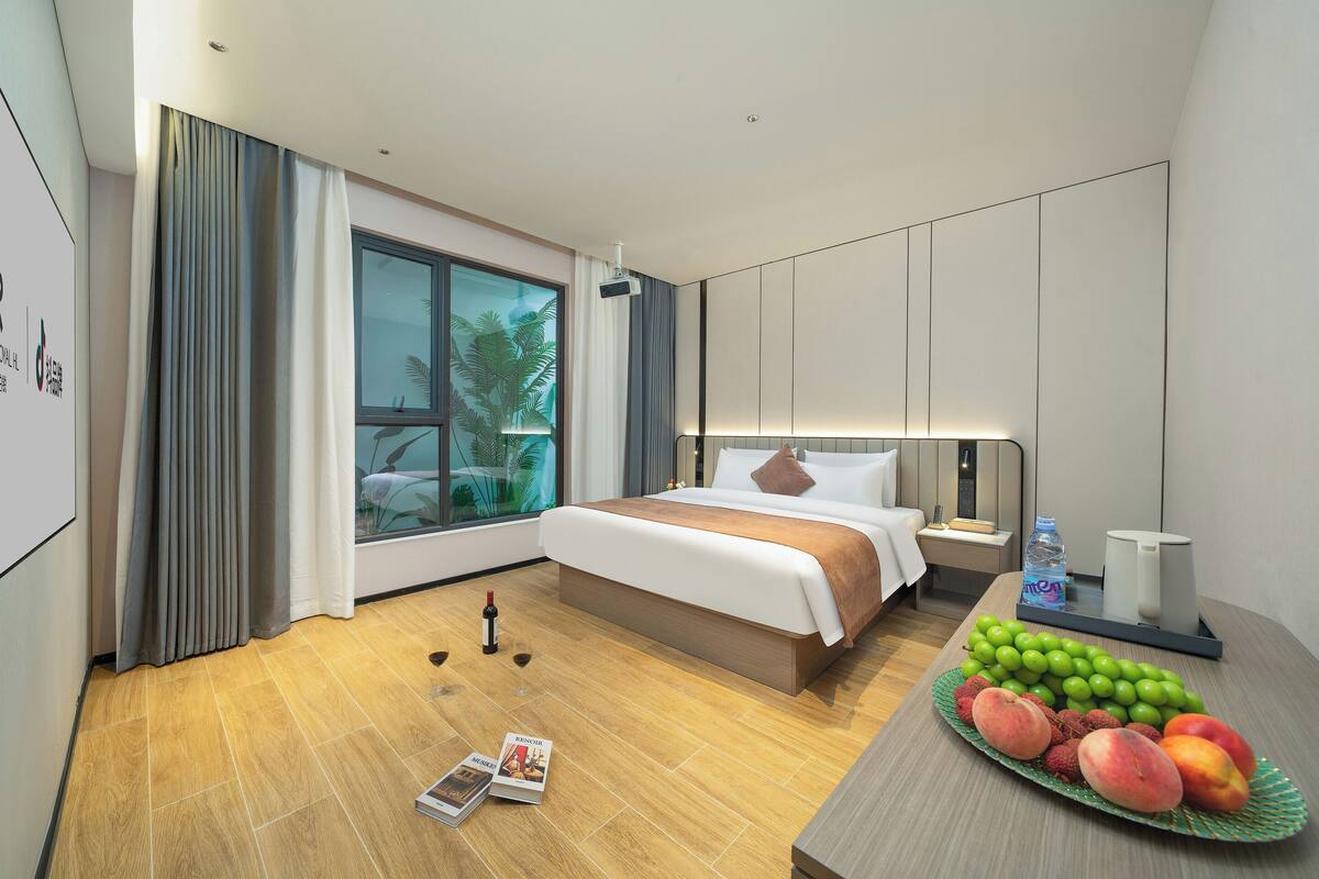 Una habitación de hotel moderna con una cama grande, una mesita de noche con una botella de agua, una bandeja de frutas y una revista en el suelo.