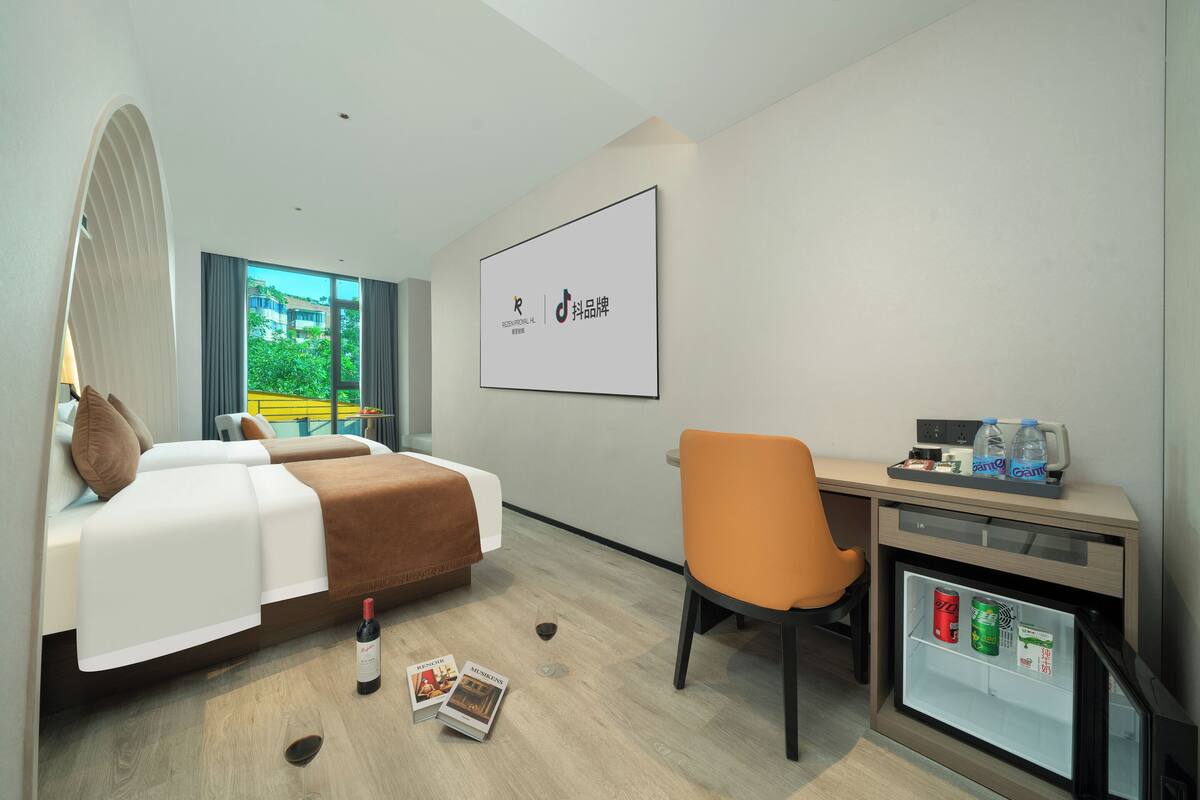 Habitación de hotel con cama, escritorio, silla, refrigerador pequeño, televisor y ventana con vista a follaje.