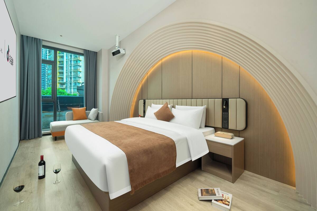 Una habitación de hotel moderna con una cama grande, una mesita de noche y un amplio ventanal con cortinas, ofreciendo vistas a un edificio alto.
