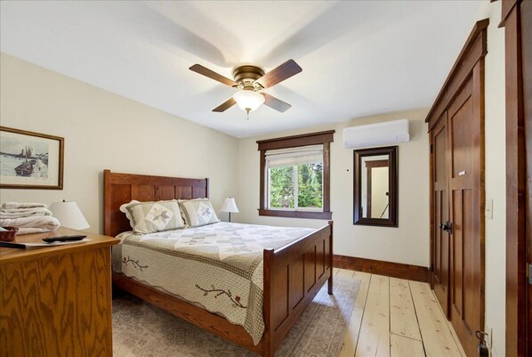 A bedroom with a wooden bed, a ceiling fan, a window with blinds, a bedside table with a lamp, and a framed picture on the wall.