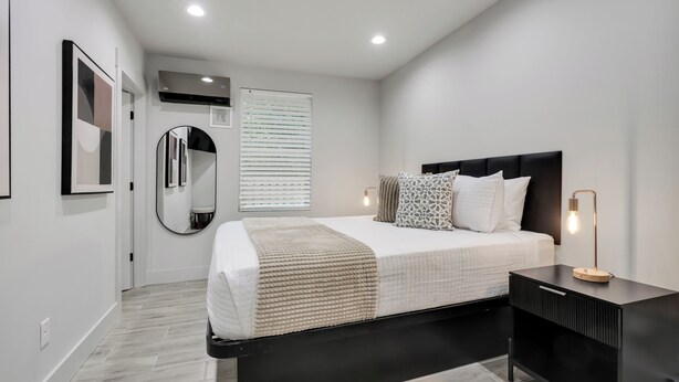 A modern bedroom with a bed, bedside tables, a mirror, and a window with blinds. - The Jewel – Modern Retreat Near YMCA & Downtown (Wilmington)