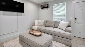 A modern living room with a grey sectional sofa, a flat-screen TV mounted on the wall, and a window with blinds. - The Jewel – Modern Retreat Near YMCA & Downtown (Wilmington)